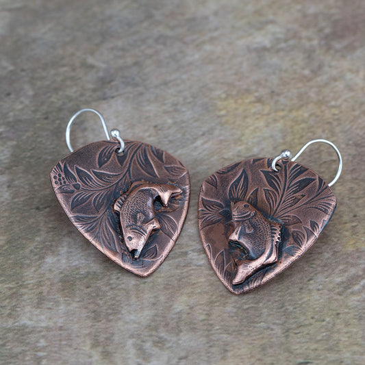 Handcrafted copper earrings featuring detailed jumping bass fish on a botanical-etched background – rustic nature-inspired fishing jewelry by Divella Designs, displayed on earthy surface.