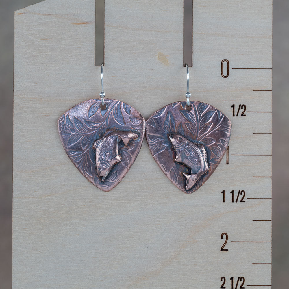 Handcrafted copper earrings featuring detailed jumping bass fish on a botanical-etched background – rustic nature-inspired fishing jewelry by Divella Designs, displayed on a wooden ruler for scale.
