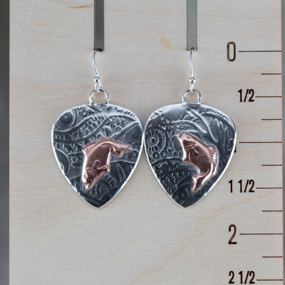 Handcrafted mixed metal earrings featuring copper jumping bass fish on textured fine silver backdrops with nature-inspired patterns – Gone Fishin’ earrings by Divella Designs, displayed on wooden ruler for scale.