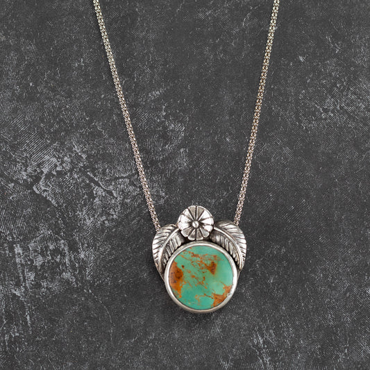 Handcrafted sterling silver pendant with round Kingman turquoise stone, accented by stamped flower and leaf details – artisan Southwestern-inspired necklace by Divella Designs, displayed on dark textured background.
