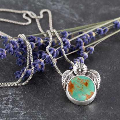 Handcrafted sterling silver pendant with round Kingman turquoise stone, accented by stamped flower and leaf details – artisan Southwestern-inspired necklace by Divella Designs, displayed on dark textured background with lilac sprigs.