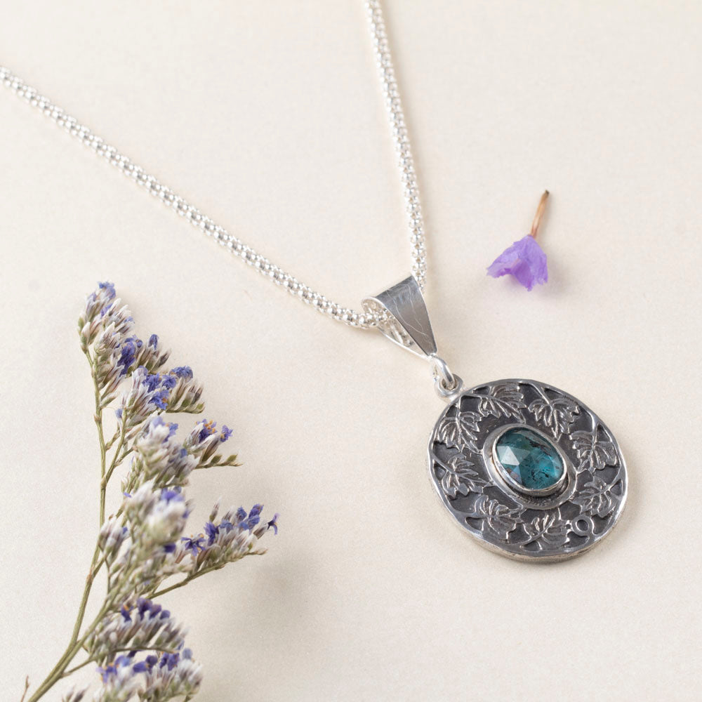 Sterling silver pendant necklace with an ornate leaf design surrounding an oval blue-green kyanite stone, shown on a white background with scattered purple flower petals