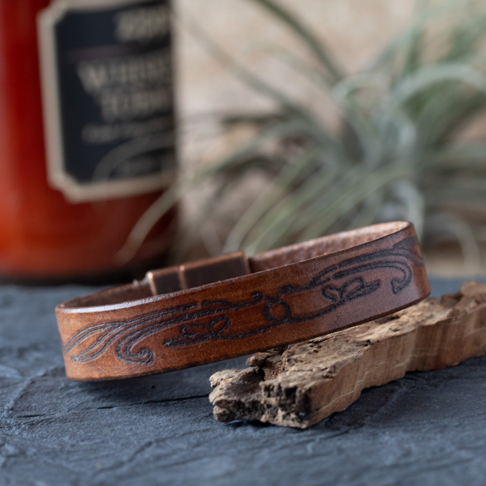 Brown leather bracelet with embossed design on a textured surface with a blurred bottle in the background
