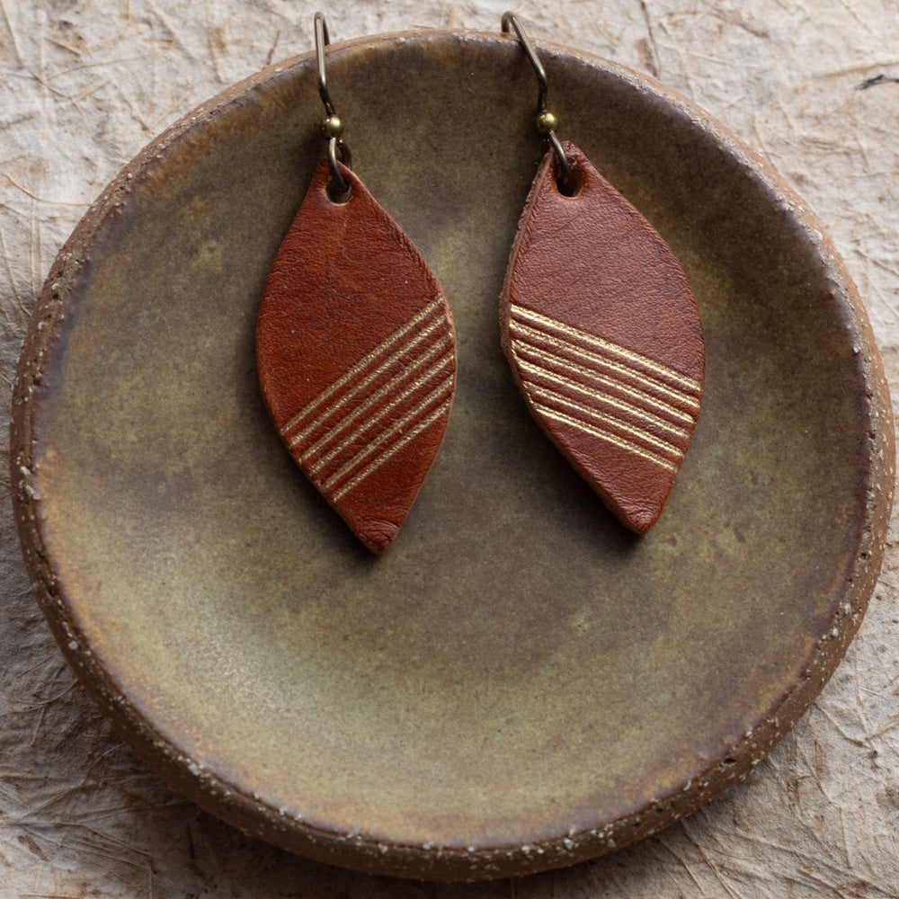 A pair of brown leather teardrop dangle earrings with gold-toned horizontal stripes, displayed on a rustic round ceramic dish.