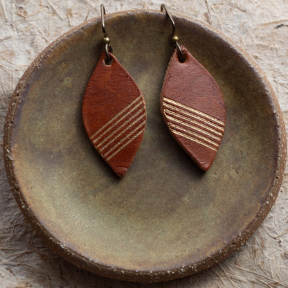 A pair of brown leather teardrop dangle earrings with gold-toned horizontal stripes, displayed on a rustic round ceramic dish.