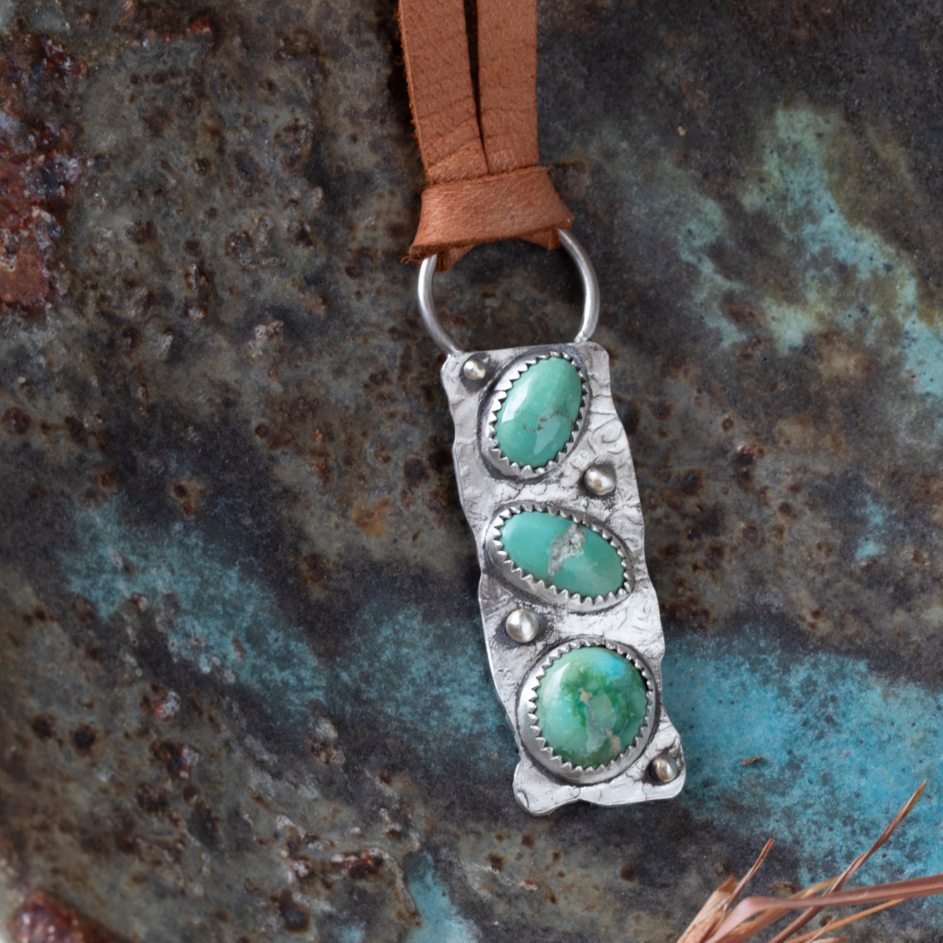 Silver pendant with turquoise stones on a brown leather strap against a textured metal background