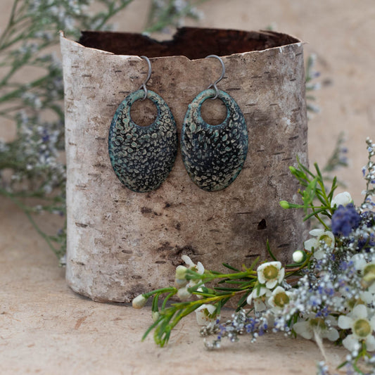 Oval-shaped enamel earrings in earthy green and black tones with a lichen-like pattern, displayed on a birch bark stand with white and purple wildflowers, handmade by Divella Designs.