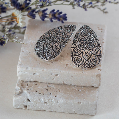 Artisan fine silver teardrop earrings with intricate mandala design, resting on natural stone with dried lavender and wildflowers – handcrafted by Divella Designs.