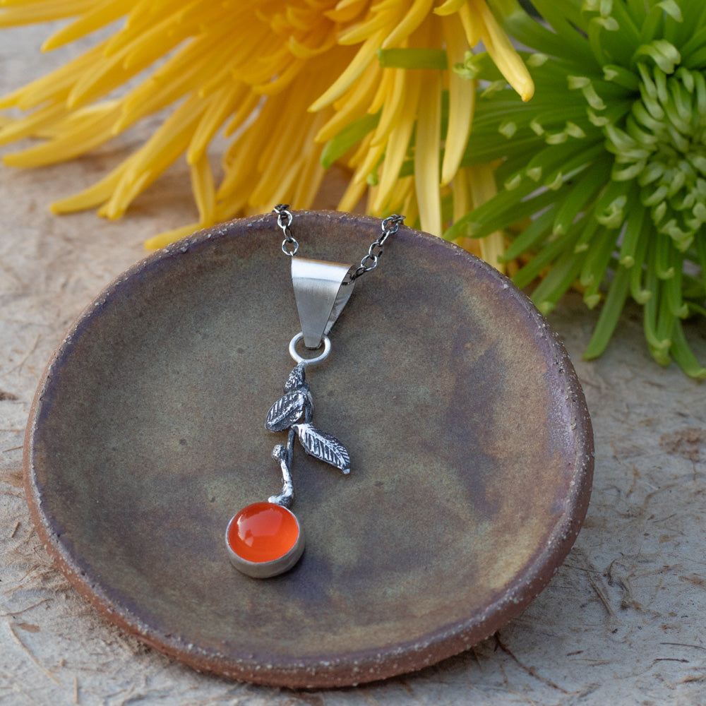 A silver pendant necklace with a round carnelian cabochon stone and silver leaves displayed on a rustic brown ceramic dish with yellow and green flowers in the background.