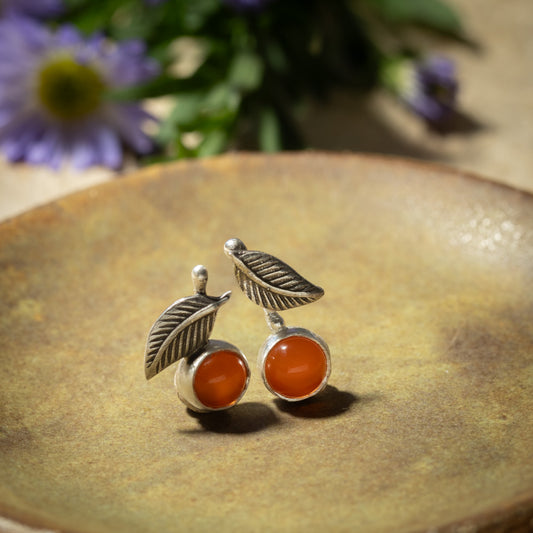 A pair of silver leaf stud earrings with round orange carnelian stones, displayed on a textured brown ceramic plate with blurred purple flowers in the background.