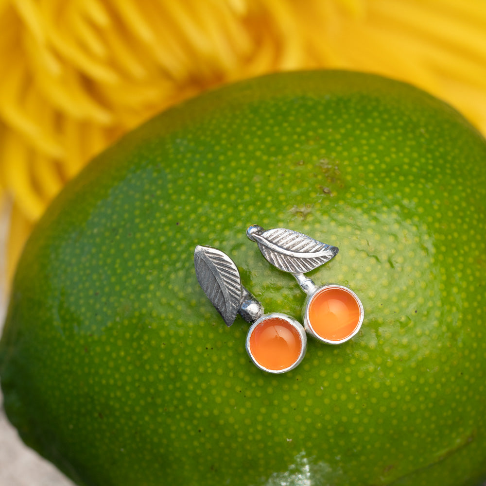 A pair of silver leaf stud earrings with orange carnelian stones, displayed on the textured green surface of a lime with a yellow background.