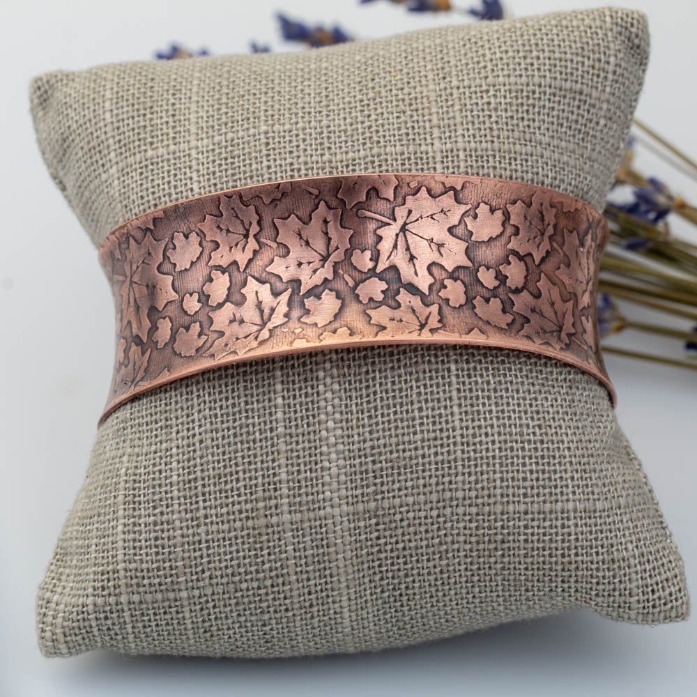 Handmade copper maple leaf cuff bracelet displayed on linen pillow.
