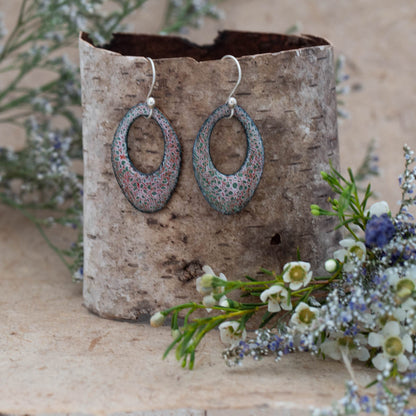 Handmade oval enamel earrings in soft green, pink, and lavender tones with a delicate lace-like pattern, displayed on a birch bark stand with wildflowers, by Divella Designs.