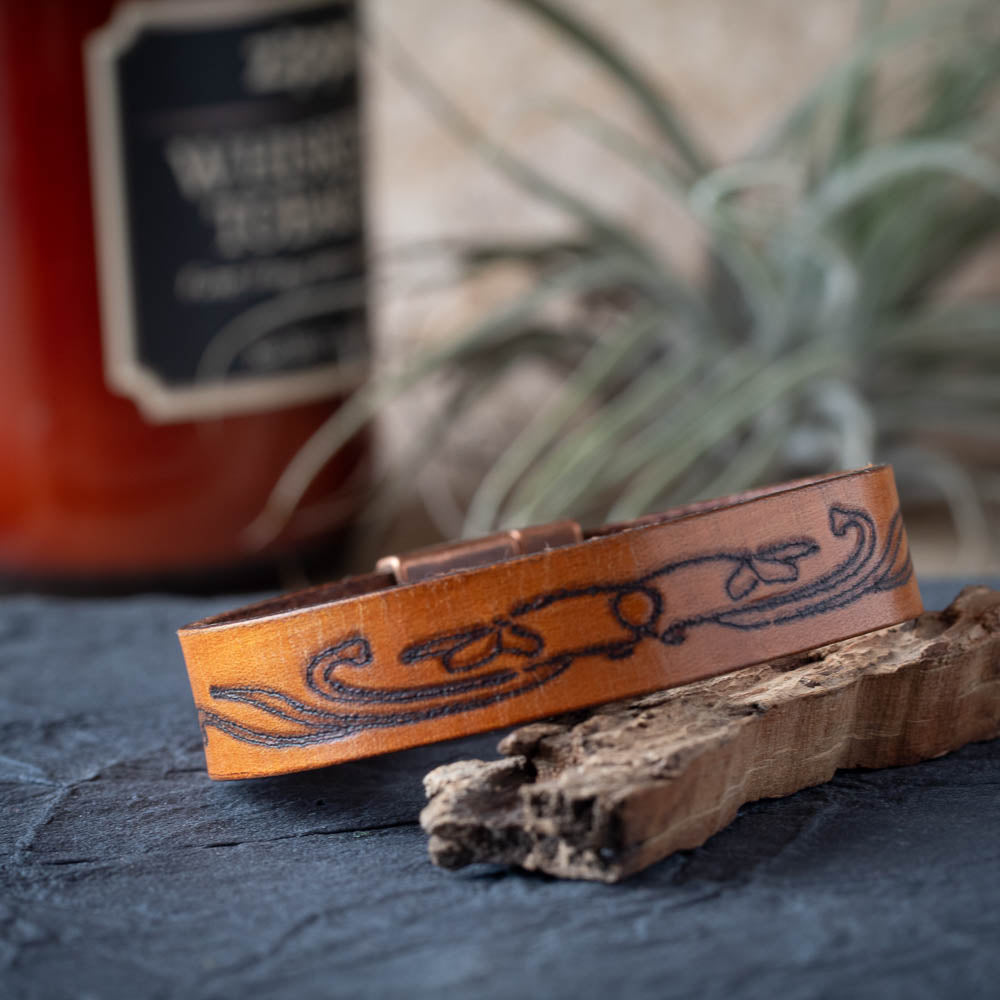 Brown leather bracelet with engraved design on a dark surface with a blurred bottle in the background