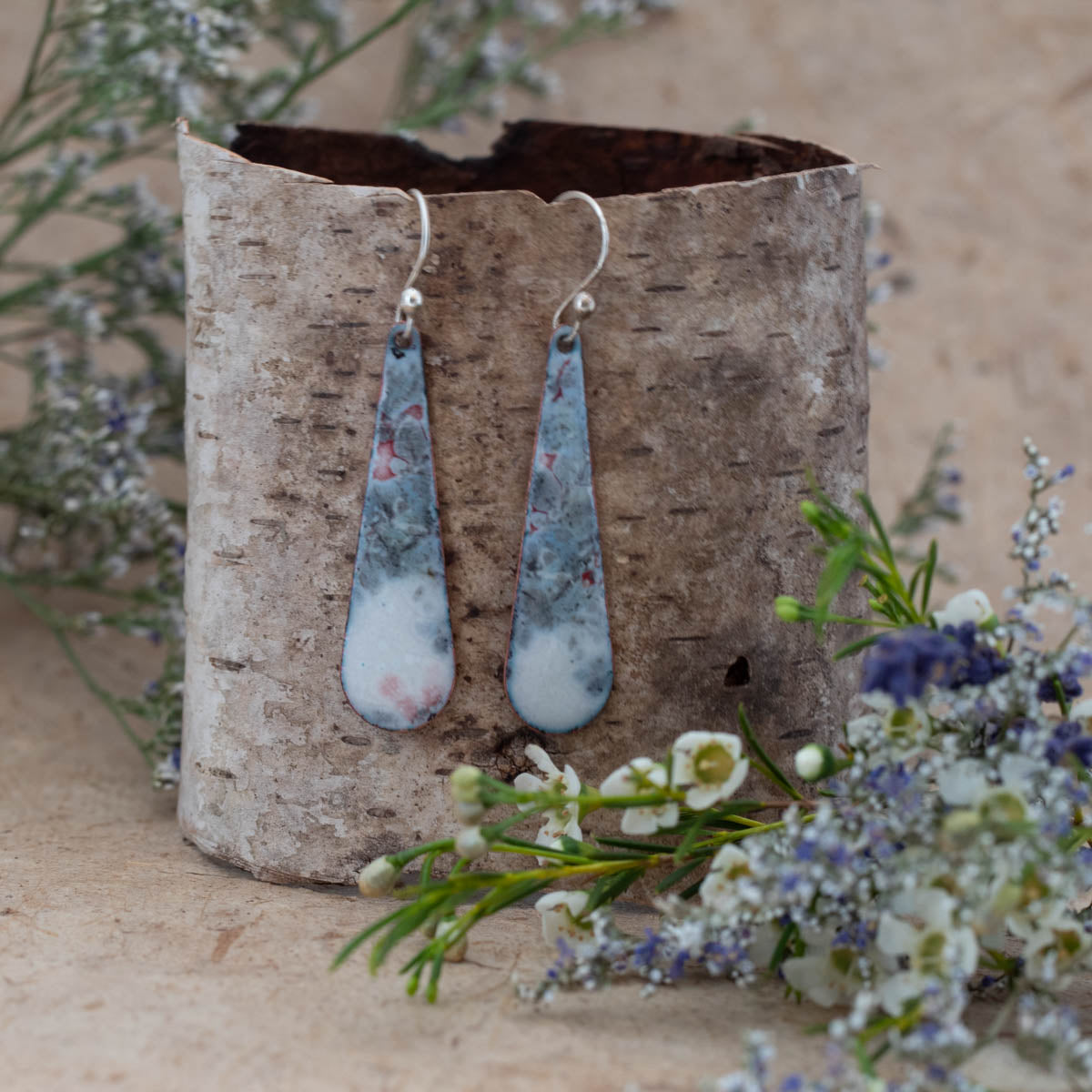 "Handcrafted enamel drop earrings in a misty blue, white, and soft pink marbled pattern, inspired by foggy forest mornings. Long teardrop shape with sterling silver ear wires, displayed on birch bark with white and lavender wildflowers – Miststone Earrings by Divella Designs.