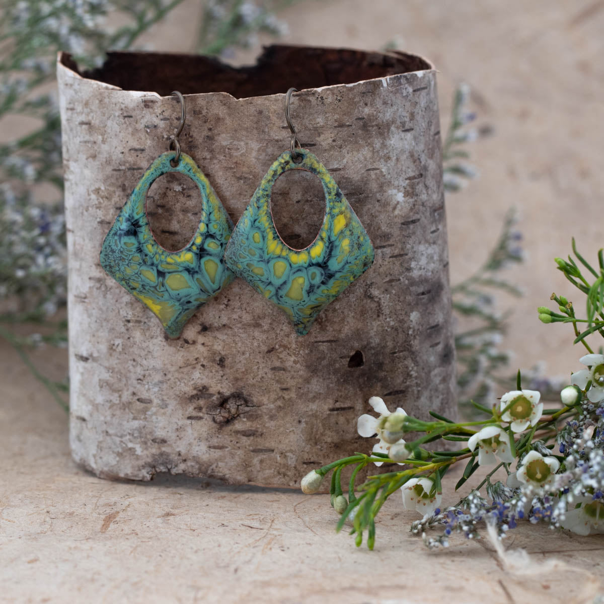 Handmade diamond-shaped enamel earrings with earthy green, yellow, and teal tones in a swirling pattern. Open oval center design with rustic patina, hanging on dark metal ear wires. Displayed on birch bark with wildflowers – Moonlight Earrings by Divella Designs.