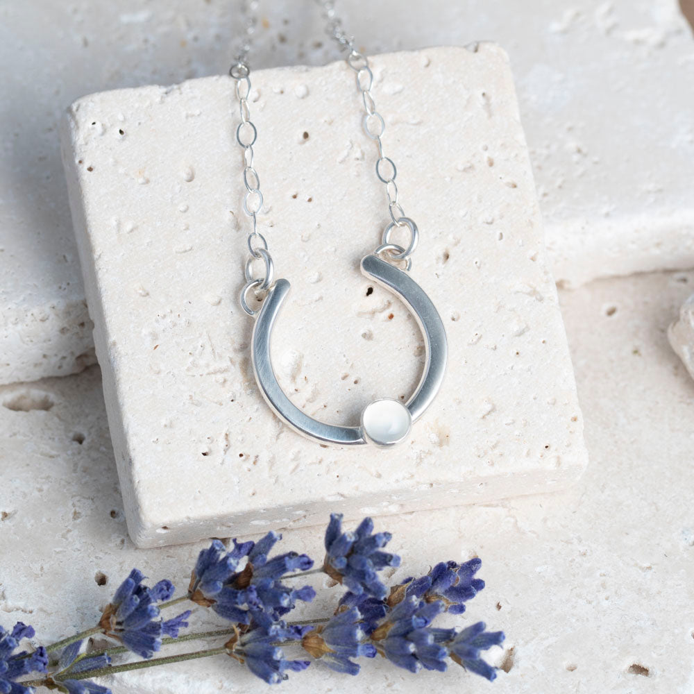 Silver necklace with a half-circle pendant set with a moonstone on a white stone surface with lavender .flowers.