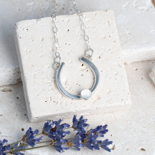 Silver necklace with a half-circle pendant set with a moonstone on a white stone surface with lavender .flowers.