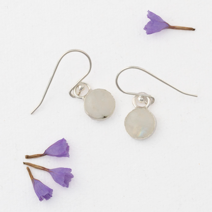 Delicate moonstone drop earrings in sterling silver with round cabochon stones, simple handmade dangles displayed with scattered purple flower petals on white background
