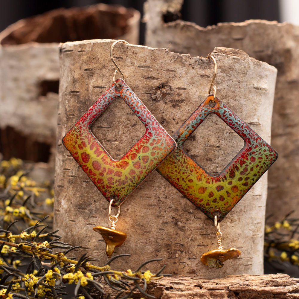 Handmade square enamel earrings in earthy red, green, and gold crackle glaze with keshi pearl dangles, displayed against birch bark and yellow wildflowers.