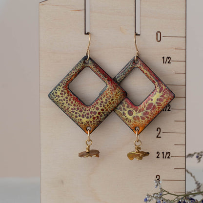 
Handmade square enamel earrings in earthy red, green, and gold crackle glaze with keishi pearl dangles dangles, displayed on a wooden ruler for scale.