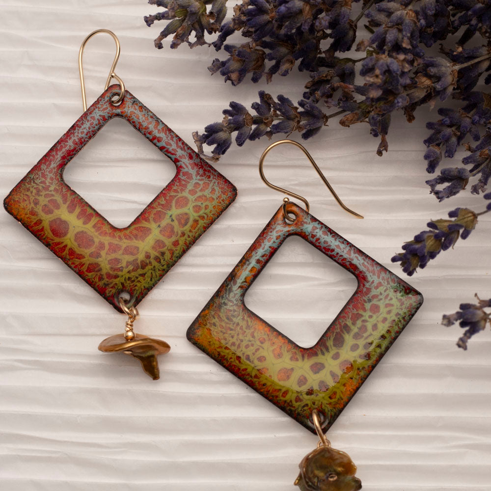 Handmade square enamel earrings in earthy red, green, and gold crackle glaze with keshi pearl dangles, displayed against white textured paper with lavender sprigs.