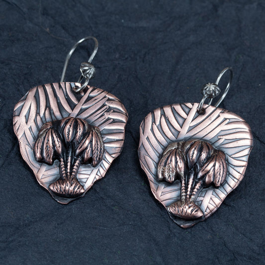 Handcrafted copper earrings with sculpted palm trees layered over embossed leaf-patterned backdrops, tropical artisan jewelry by Divella Designs – displayed on dark slate background.