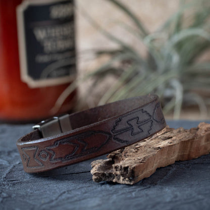Brown leather bracelet with embossed design on a textured surface with a blurred bottle in the background