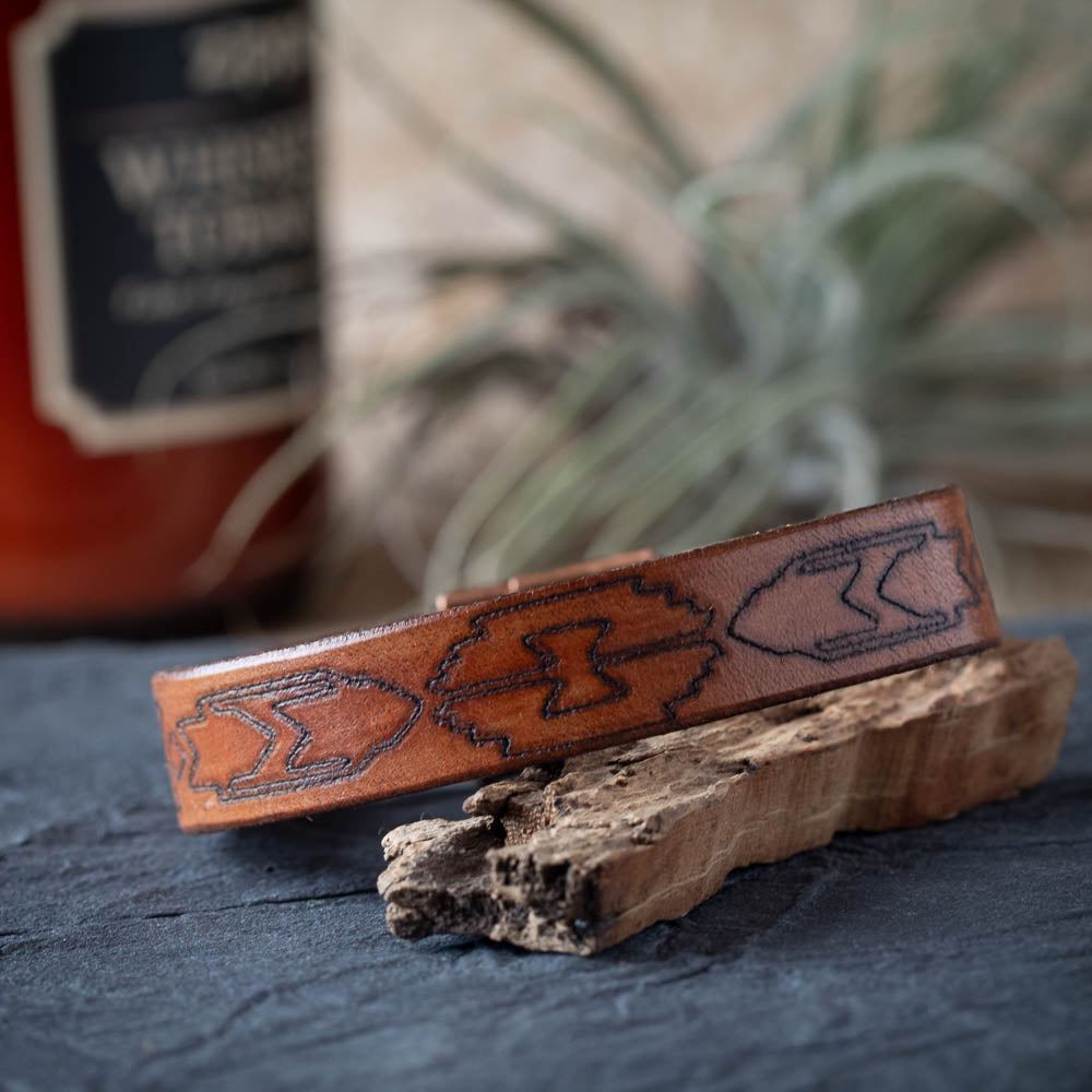 Brown leather cuff with embossed design on a dark surface with a blurred bottle and plant in the background