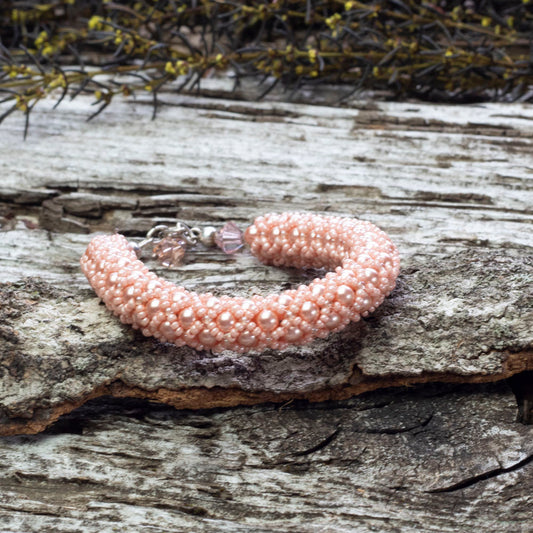 Peach rose beaded bangle bracelet made with Swarovski pearls, displayed on rustic weathered wood with sprigs of greenery in the background.