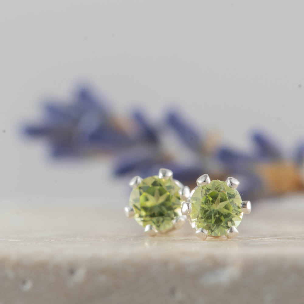Peridot gemstone stud earrings on a light surface with blurred flowers in the background