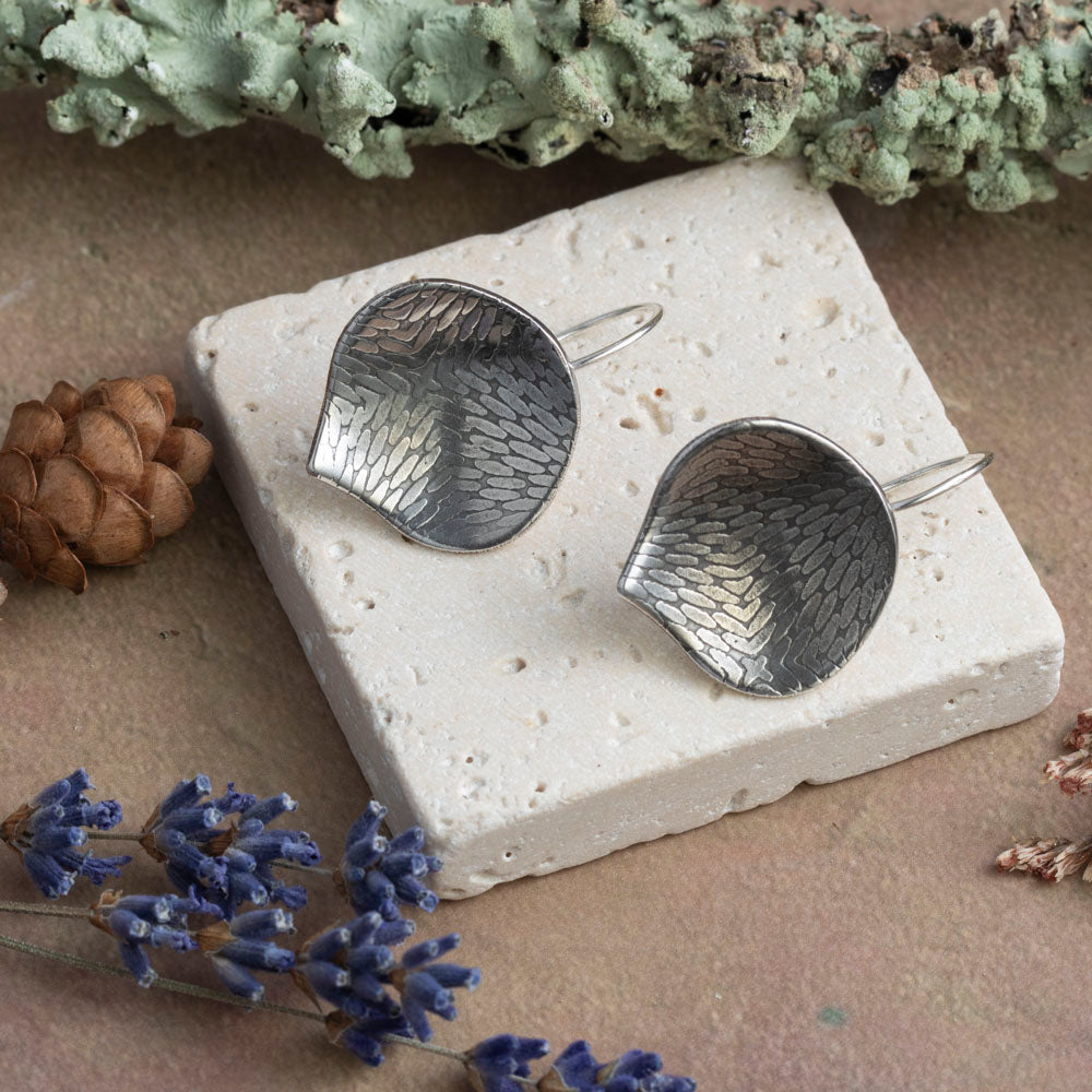 Silver leaf-shaped earrings on a stone surface with natural elements like moss, pine cone, and lavender.