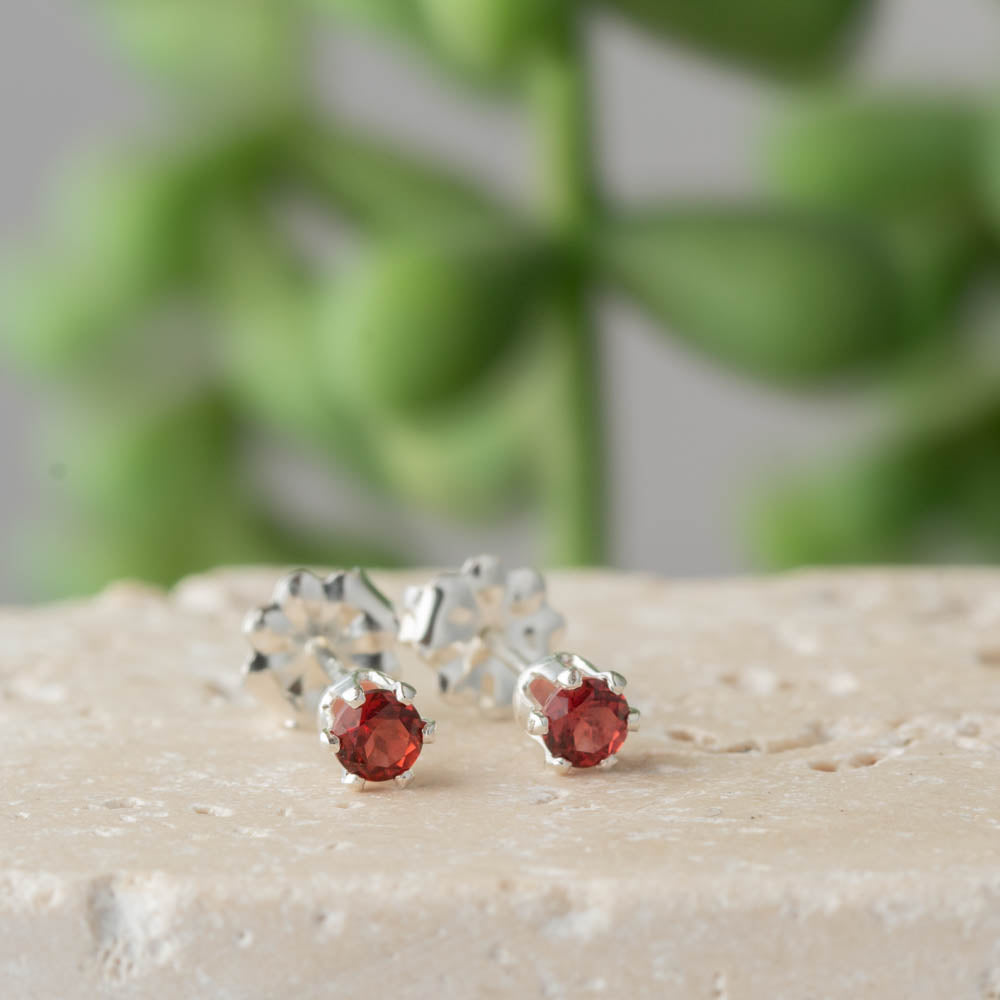 Silver earrings with red garnet gemstones on a beige surface with a blurred green plant background