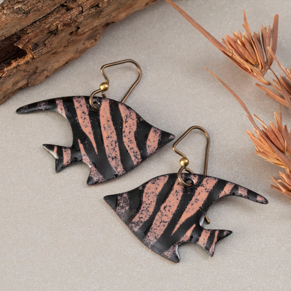 Handmade enamel fish-shaped earrings in bold pink and black stripes, styled with driftwood and natural textures – artisan jewelry by Divella Designs