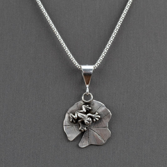 Handcrafted sterling silver frog pendant resting on a textured lily pad, nature-inspired Pond Jumper necklace by Divella Designs – shown on gray background with popcorn chain.