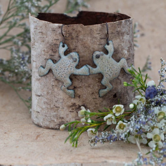 Whimsical handmade enamel earrings shaped like playful frogs, finished in a soft aqua and ivory crackle glaze. Shown on birch bark with wildflower accents – Pond Pals Enamel Earrings by Divella Designs.