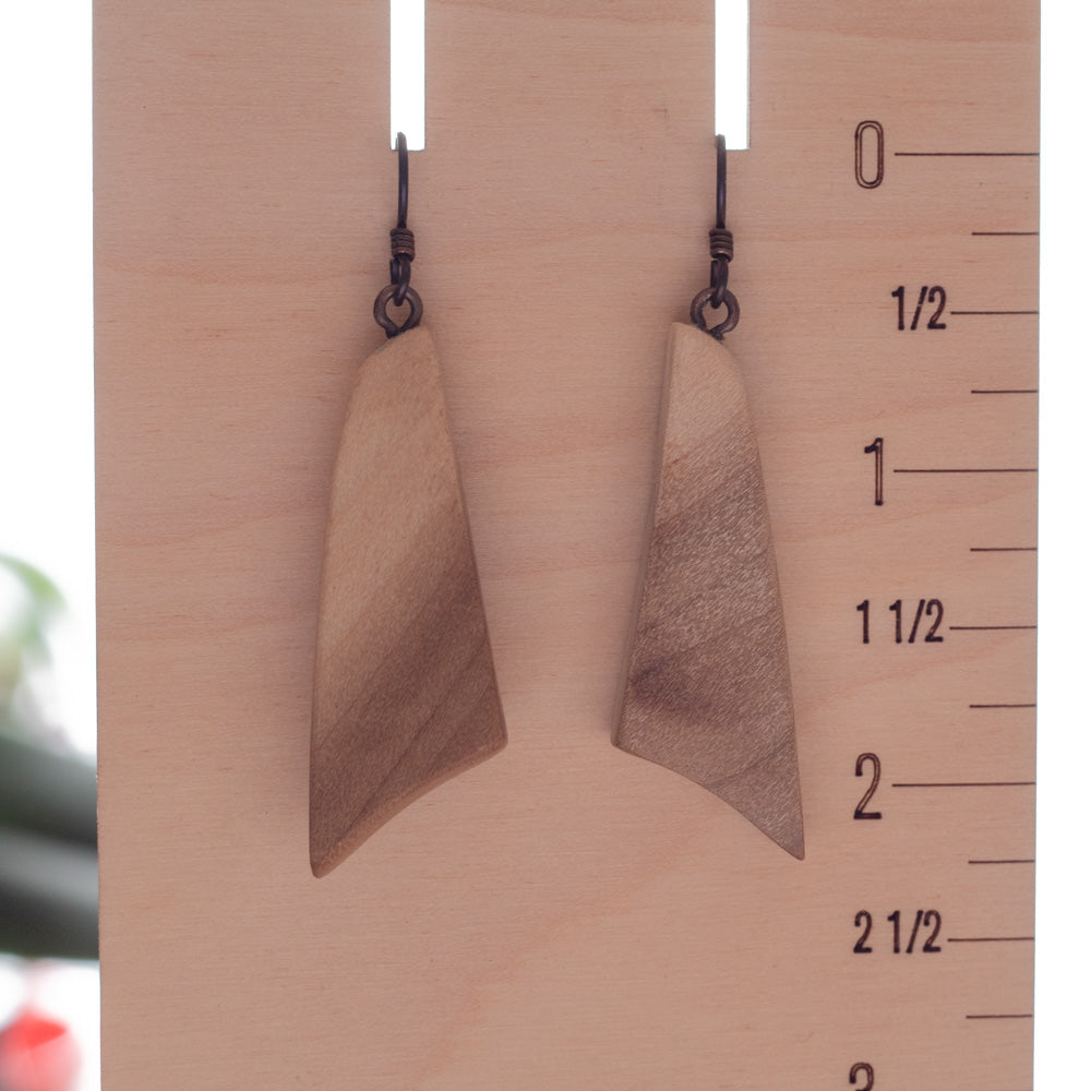 A pair of handmade wooden dangle earrings with triangular shapes and dark metal hooks, displayed on a wooden block with measurement markings.