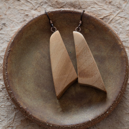 A pair of wooden drop earrings with elongated triangular shapes, displayed on a round rustic ceramic dish over a textured beige surface.
