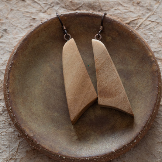 A pair of wooden drop earrings with elongated triangular shapes, displayed on a round rustic ceramic dish over a textured beige surface.