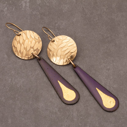 Purple enamel teardrop earrings featuring 24k gold droplet accents and round hammered brass discs, displayed on a gray jewelry stand with a light background.