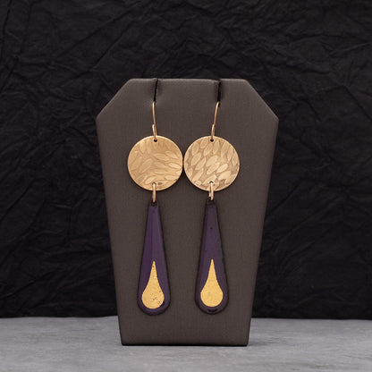 Purple enamel teardrop earrings featuring 24k gold droplet accents and round hammered brass discs, displayed on a gray jewelry stand with a dark crinkled background.