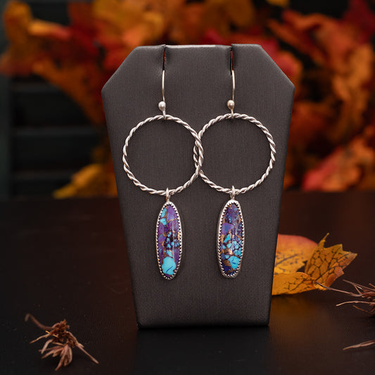 Handcrafted dangle earrings featuring oval purple Mohave turquoise and Kingman turquoise stones set in sterling silver with twisted silver hoops, displayed against an autumn background.
