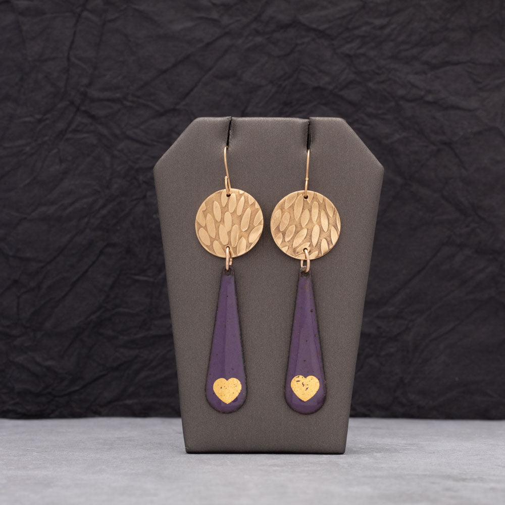 Handmade purple enamel teardrop earrings with 24k gold heart accents and hammered brass discs, displayed on a gray earring stand with dark textured background.
