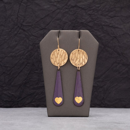 Handmade purple enamel teardrop earrings with 24k gold heart accents and hammered brass discs, displayed on a gray earring stand with dark textured background.