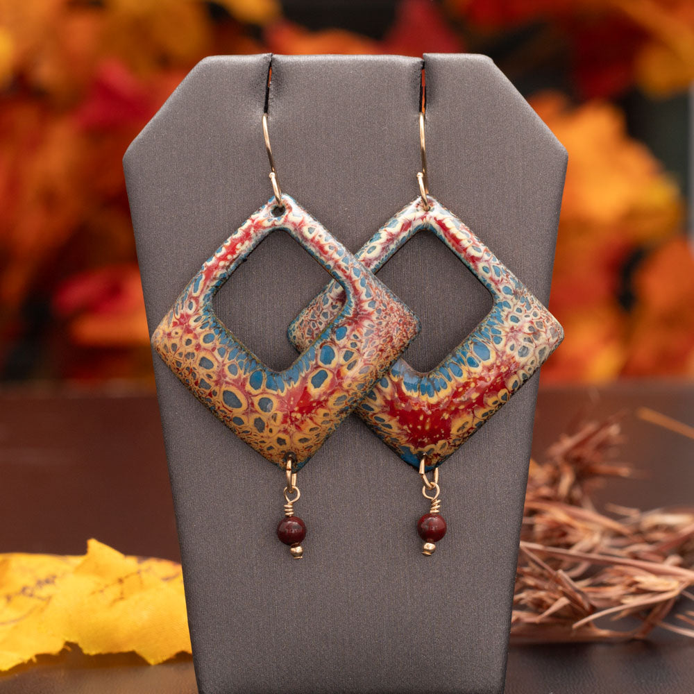 Square-cut enamel earrings in warm red, gold, and blue tones with a speckled pattern and small round red bead dangles, displayed on a black background with autumn leaves nearby.