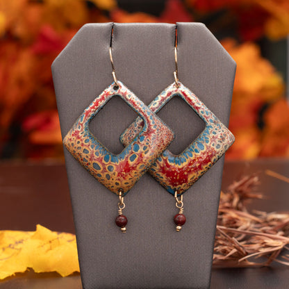 Square-cut enamel earrings in warm red, gold, and blue tones with a speckled pattern and small round red bead dangles, displayed on a black background with autumn leaves nearby.