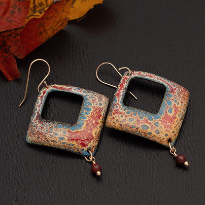 Square-cut enamel earrings in warm red, gold, and blue tones with a speckled pattern and small round red bead dangles, displayed on a black background with autumn leaves nearby.