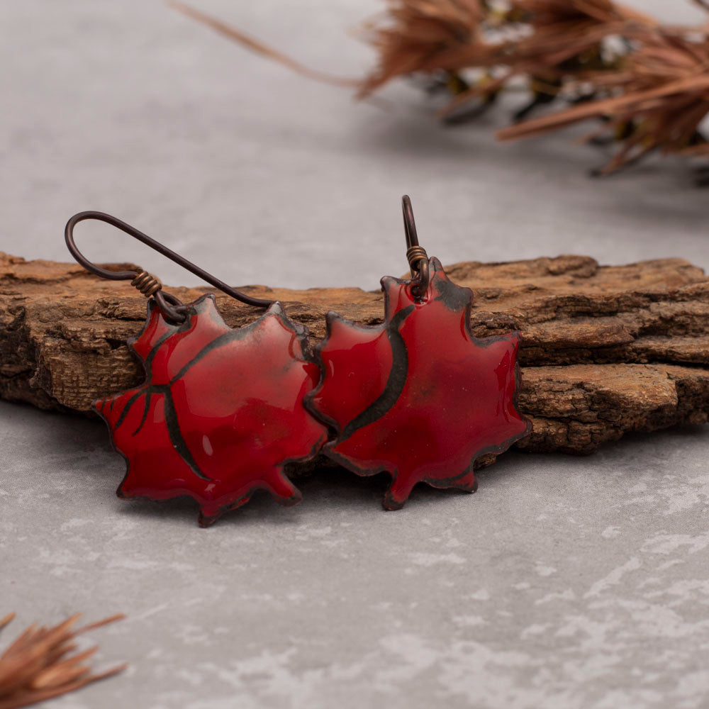 Red maple leaf-shaped earrings on a wooden branch with a neutral background
