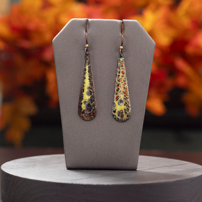 Brown, orange, and yellow enamel teardrop earrings displayed with autumn leaves on a dark background.
