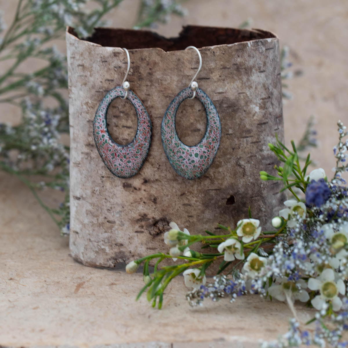 Handmade oval enamel earrings in soft green, pink, and lavender tones with a delicate lace-like pattern, displayed on a birch bark stand with wildflowers, by Divella Designs.